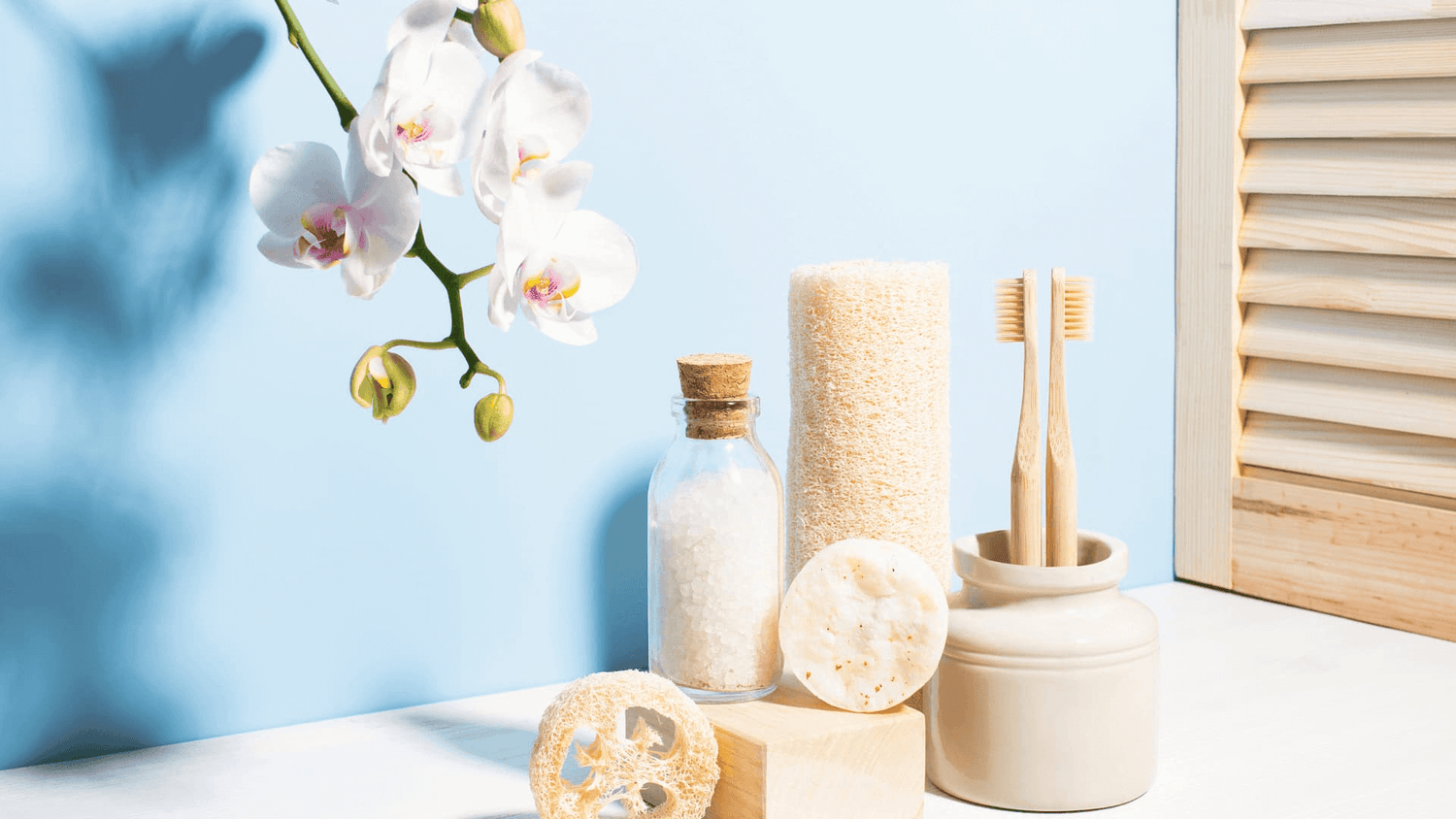Set de baño natural con esponjas, cepillos, sales y flores blancas sobre una superficie blanca, con fondo azul claro. Ambiente relajante y minimalista ideal para el autocuidado y spa en casa.