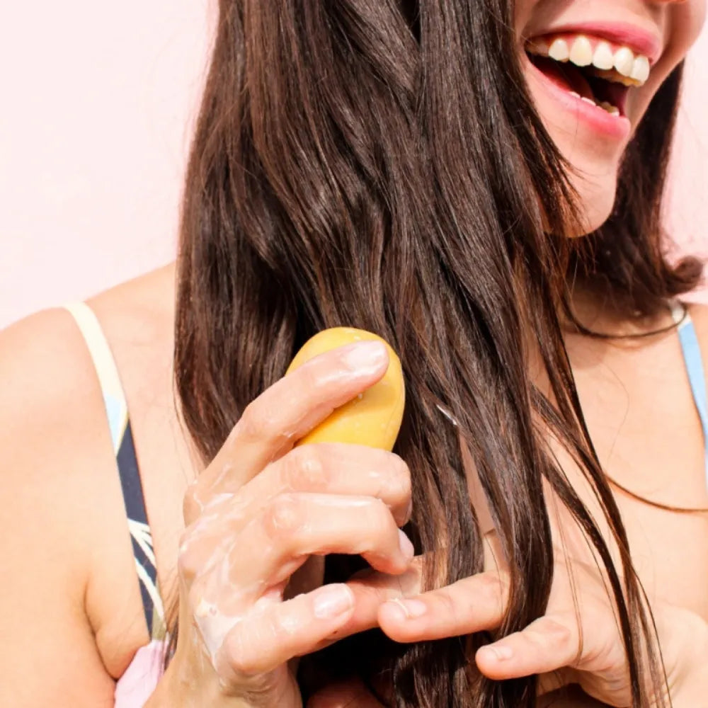 Fotografía de una mujer sonriendo mientras aplica acondicionador sólido en barra de color ámbar sobre su cabello mojado. La imagen muestra un momento de uso del producto durante la rutina de cuidado capilar.