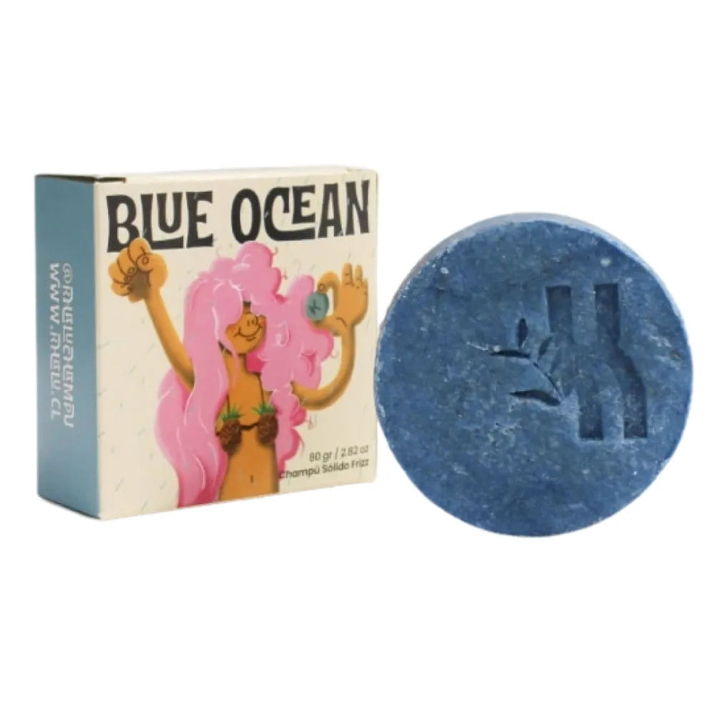 Foto de un champú sólido azul en barra con relieve de rama y letra “K”, acompañado de su empaque ilustrado con una figura femenina de cabello rosado. El producto es de la línea “Blue Ocean”, especial para el control del frizz.