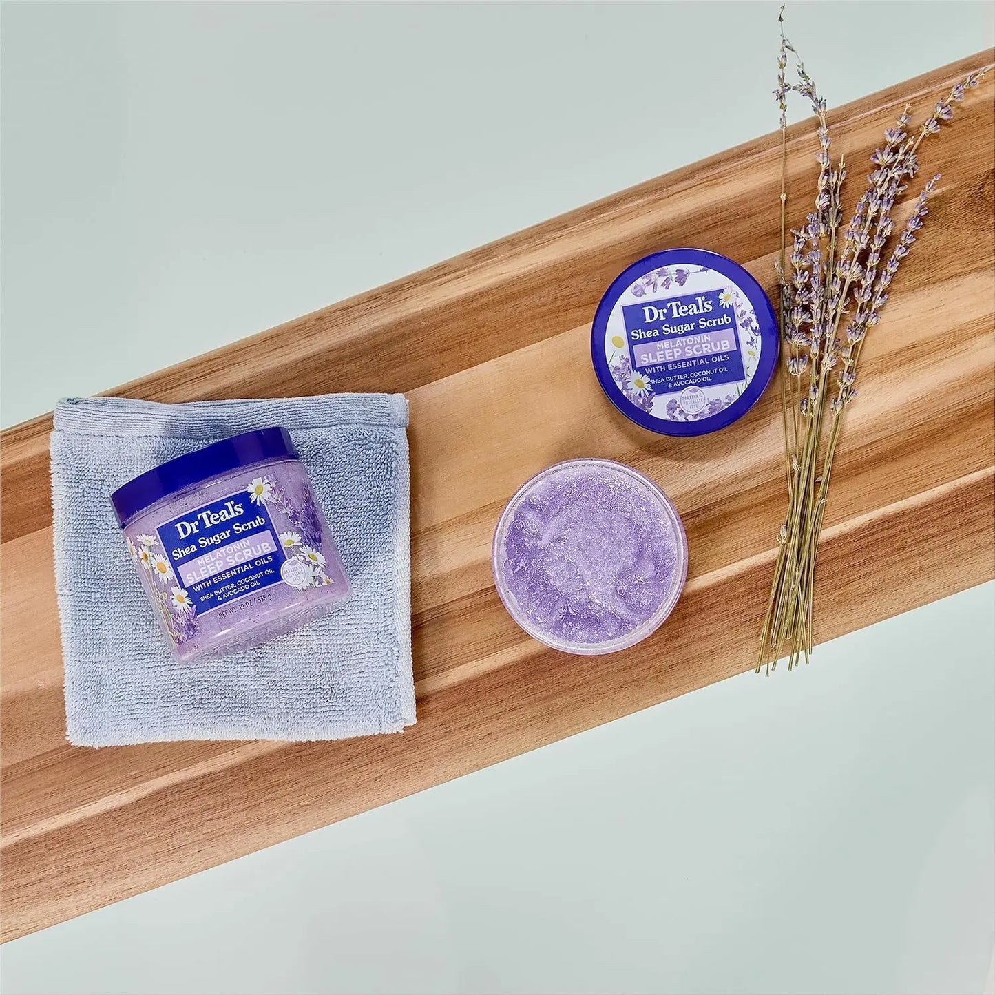 Pote de exfoliante Dr. Teal’s Sleep Scrub colocado sobre una bandeja de madera con una toalla, flores de lavanda y otro pote abierto mostrando su textura.