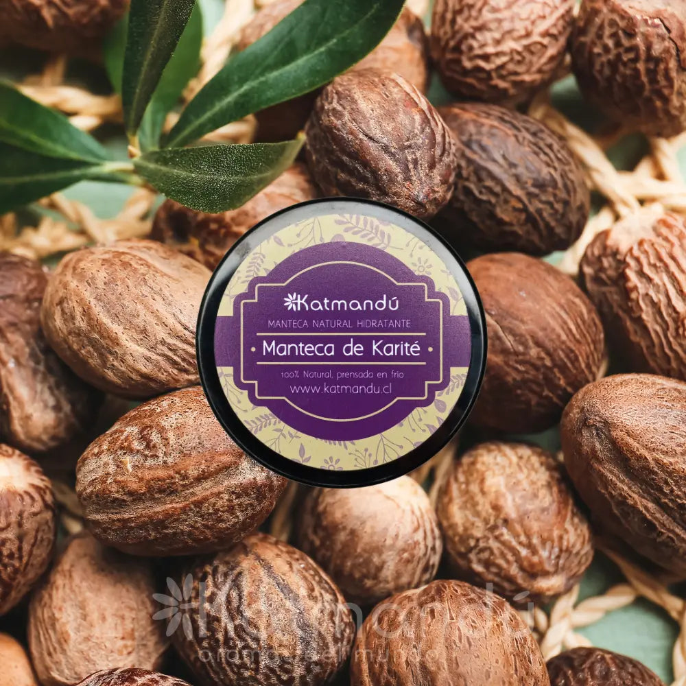 Envase de manteca de karité Katmandú sobre fondo de nueces de karité. El producto es 100% natural, prensado en frío e hidratante. La tapa morada muestra la marca, nombre del producto y sitio web. Ideal para cuidado de la piel de manera natural y nutritiva.