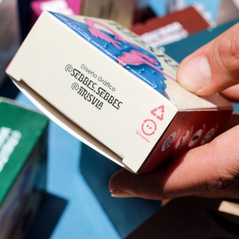 Enfoca en un costado de la caja del producto “RASSRAW”, sostenida por una mano. Se pueden ver los créditos del diseño gráfico, incluye los símbolos de reciclaje y sostenibilidad, indicando que el empaque es reciclable.