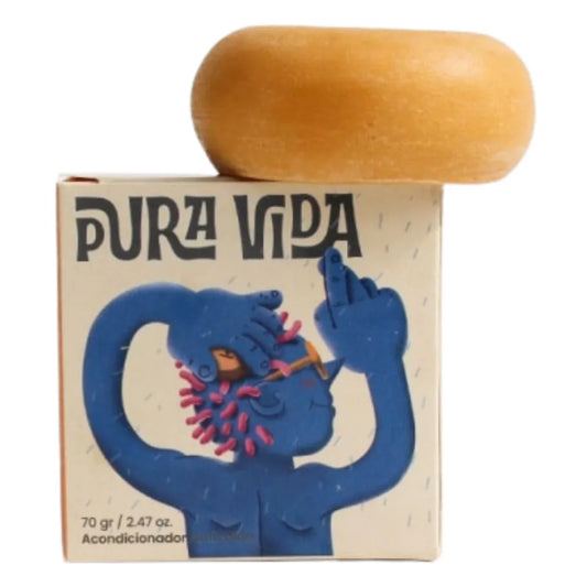 Fotografía de acondicionador sólido en barra de color ámbar, colocado sobre una caja con diseño ilustrado de persona azul con gafas y cabello rosa. La caja muestra el texto “PURA VIDA” y detalla que contiene 70 gramos de producto.
