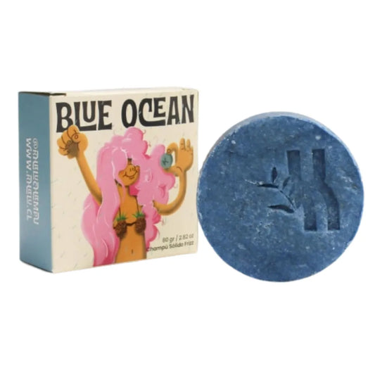 Foto de un champú sólido azul en barra con relieve de rama y letra “K”, acompañado de su empaque ilustrado con una figura femenina de cabello rosado. El producto es de la línea “Blue Ocean”, especial para el control del frizz.