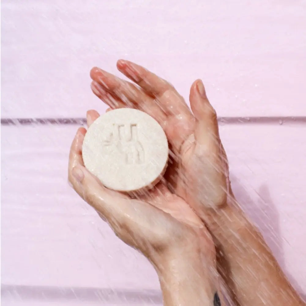 Fotografía de manos sosteniendo un champú sólido redondo bajo el agua de la ducha, con fondo rosado. El champú tiene el logotipo de la marca en relieve visible en el centro.