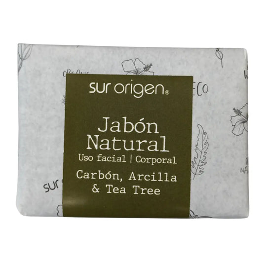 Jabón natural Sur Origen para uso facial y corporal, envuelto en papel decorado. Etiqueta verde indica sus ingredientes: Carbón, Arcilla y Tea Tree. Producto artesanal de aspecto ecológico y natural.