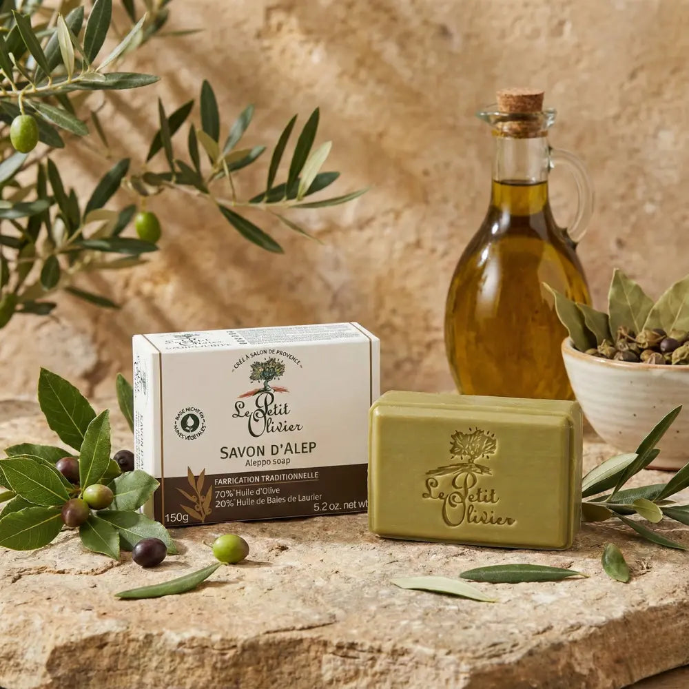 Jabón de Alepo Le Petit Olivier con caja y barra visible, acompañado de ingredientes naturales como aceite de oliva, hojas de laurel y aceitunas, sobre fondo rústico que resalta su origen artesanal y natural.