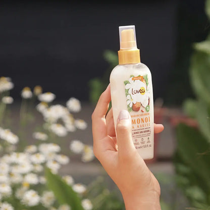 Foto del spray Lovea Monoi & Karité 150 ml sostenido con una mano al aire libre, flores blancas de fondo